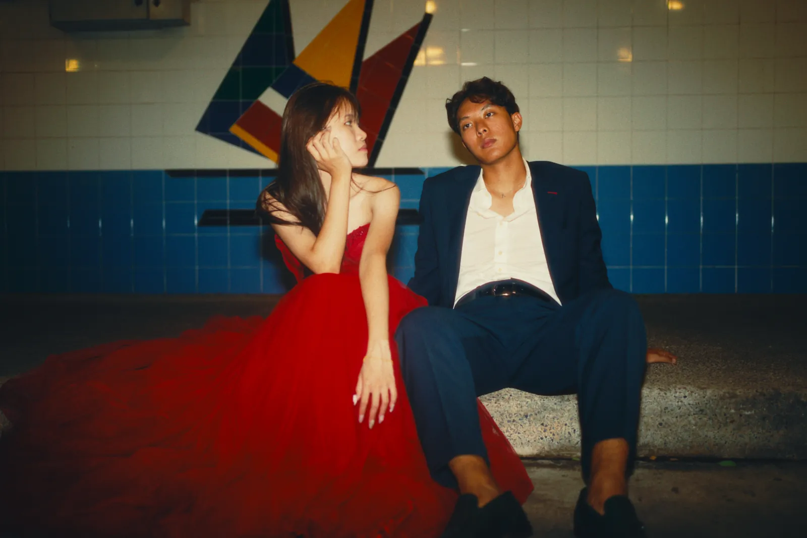Julius in a dark suit seated beside a model in a red dress against a tiled wall.