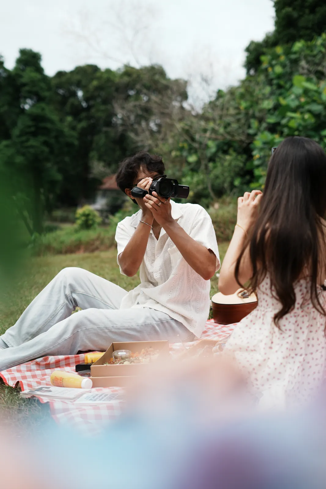 Julius in white holding a camera on a picnic blanket opposite another model.