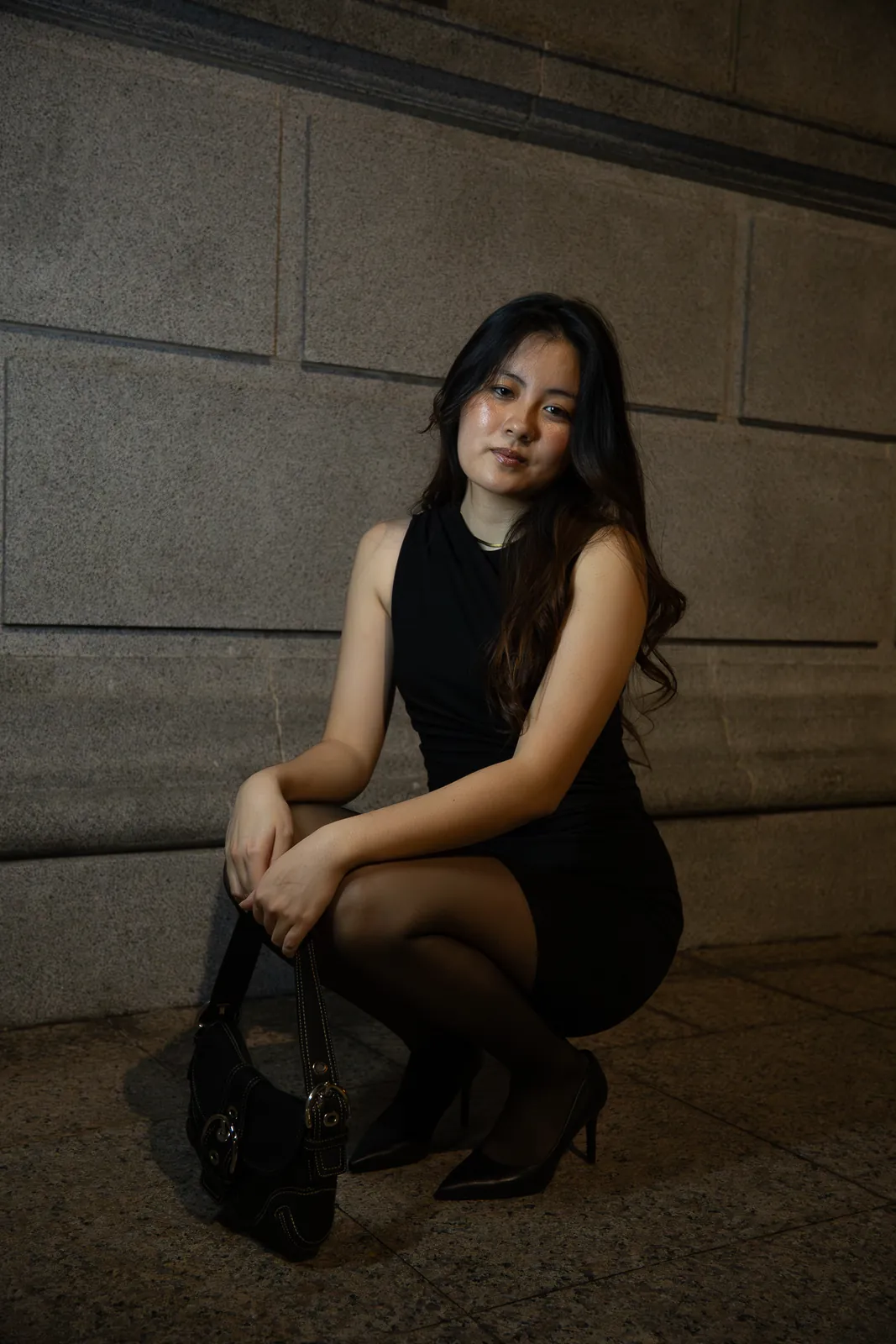 Woman in a black dress seated against a stone wall in soft shadow.