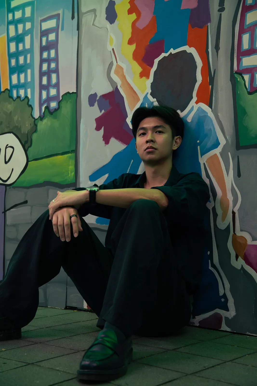 Man dressed in black seated in front of a colourful painted mural.
