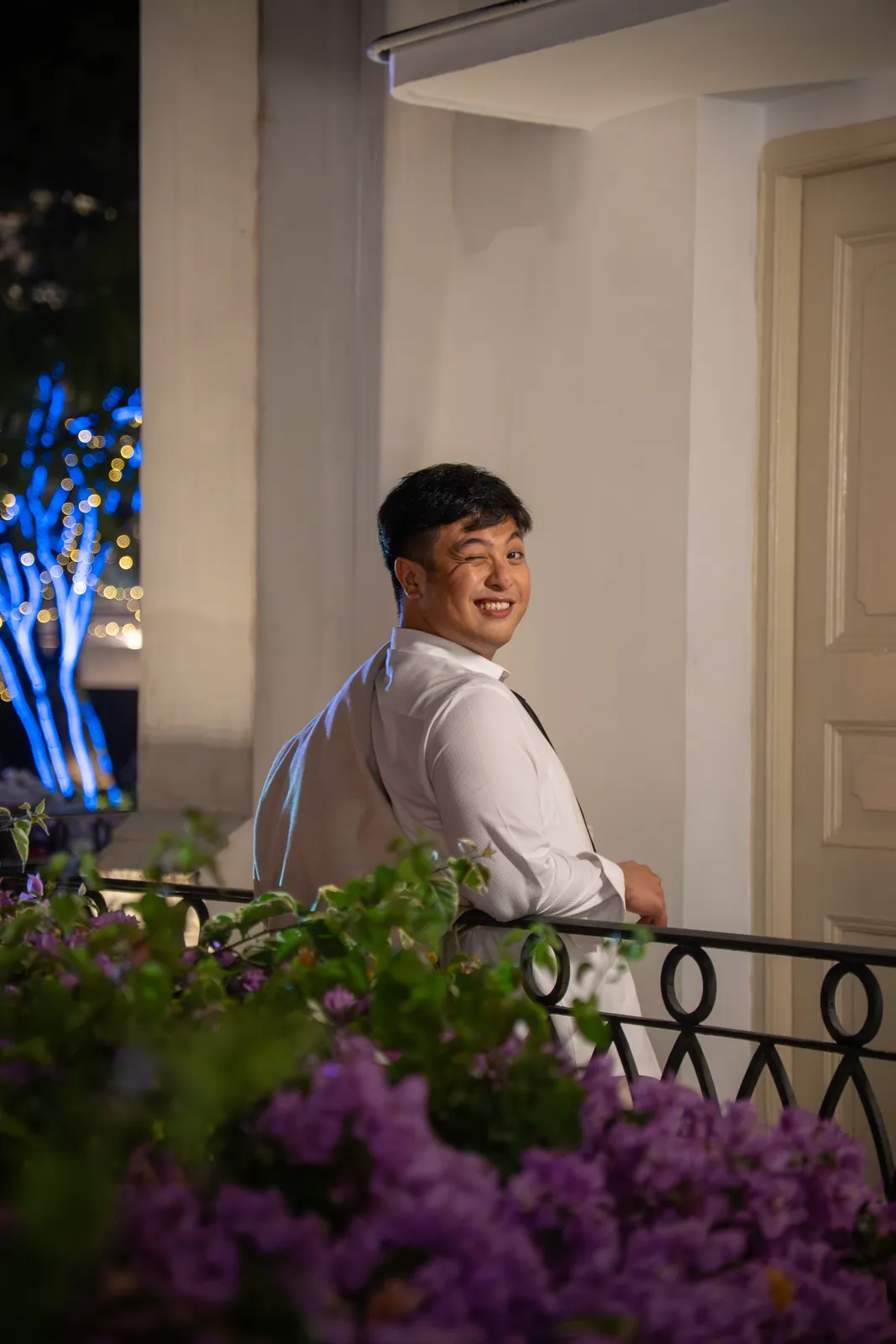 Man in a white shirt turning toward the camera above a balcony of purple flowers.