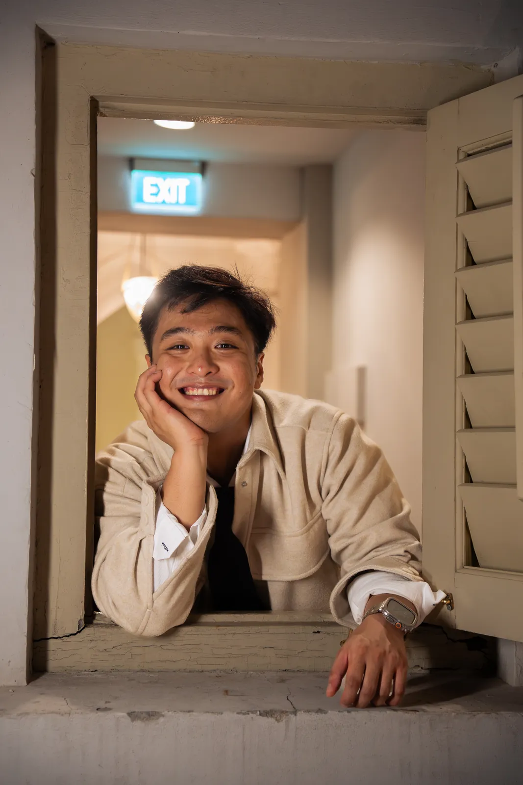 Man in a light jacket smiling through an open window beneath an exit sign.