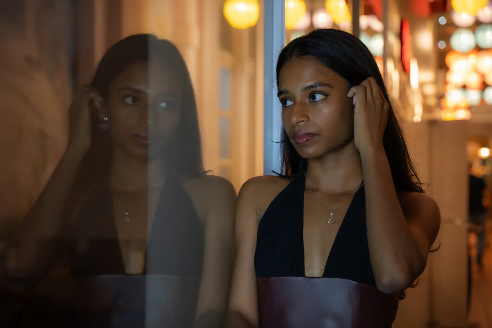 Woman beside a reflective window with her reflection and warm lights behind her.