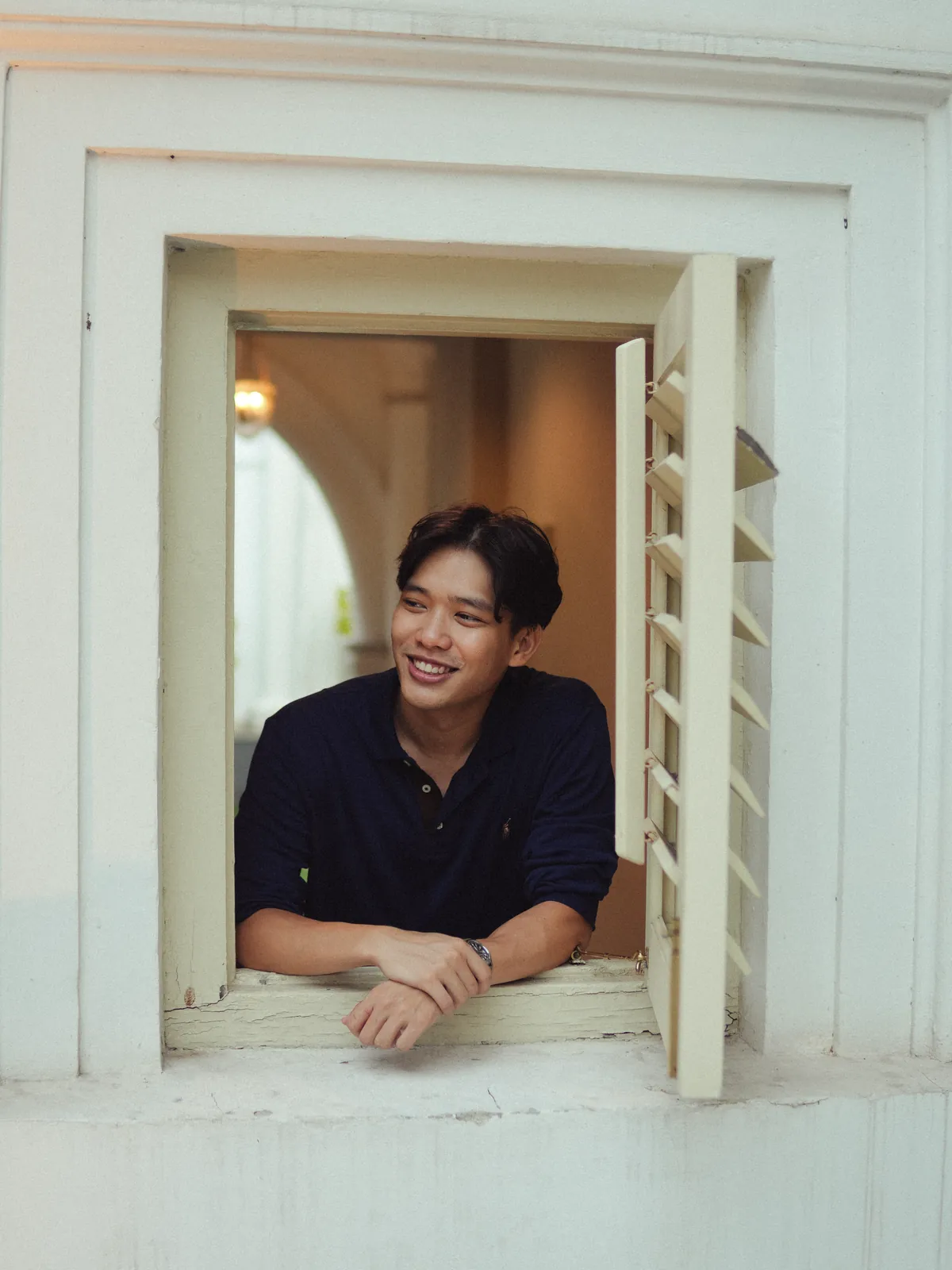 Man in a dark shirt leaning out through pale wooden window shutters.