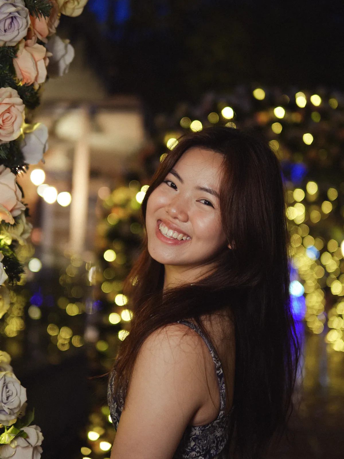 Woman smiling beside flowers with warm lights in the night-time background.