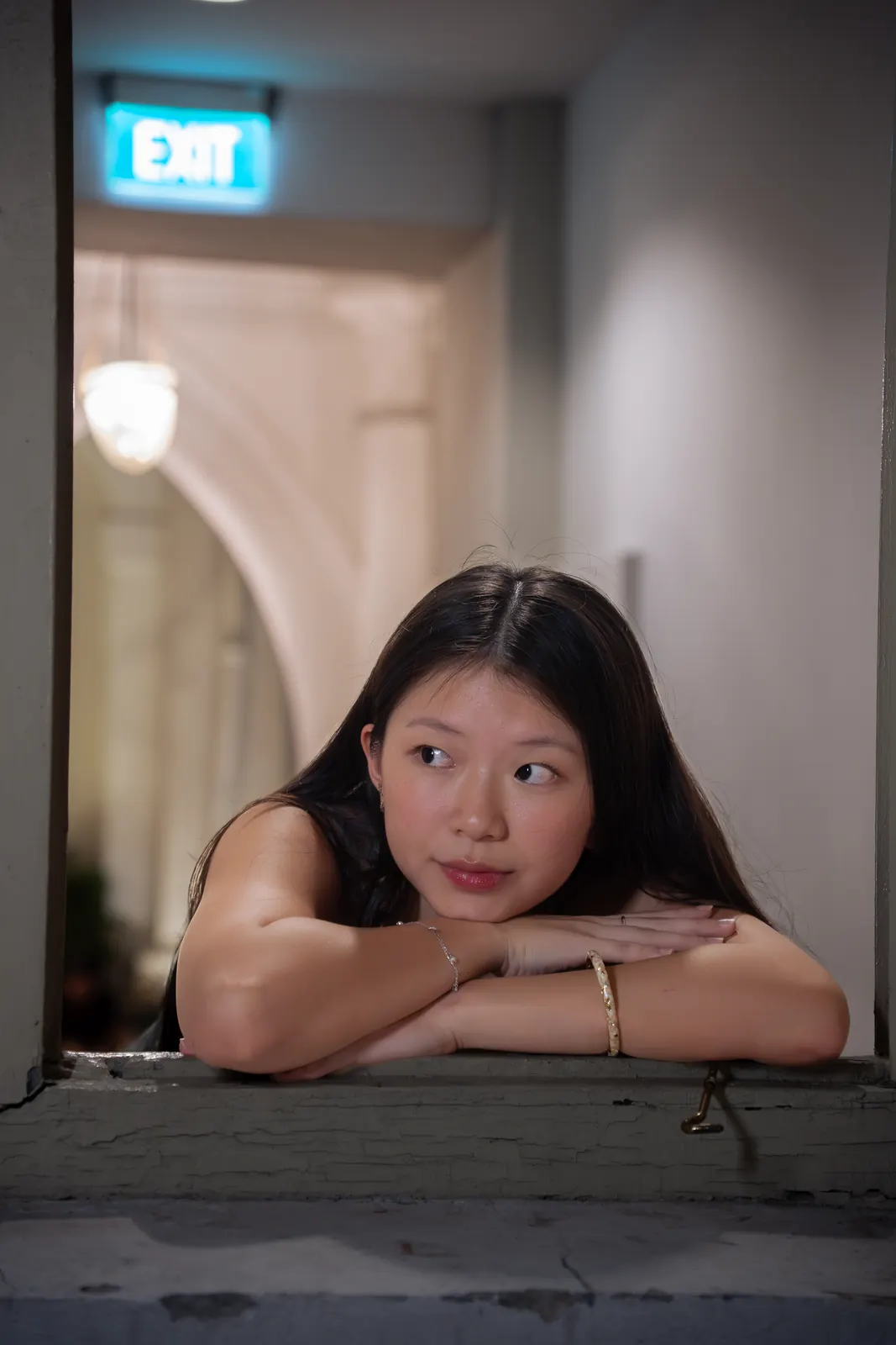 Woman leaning against a window frame beneath a red exit sign in a softly lit corridor.