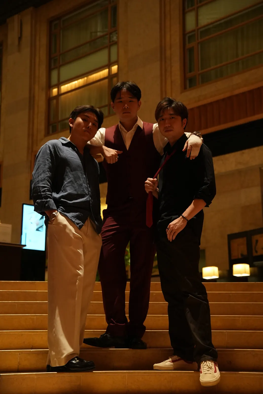 Three men posing together outside a warmly lit building at night.