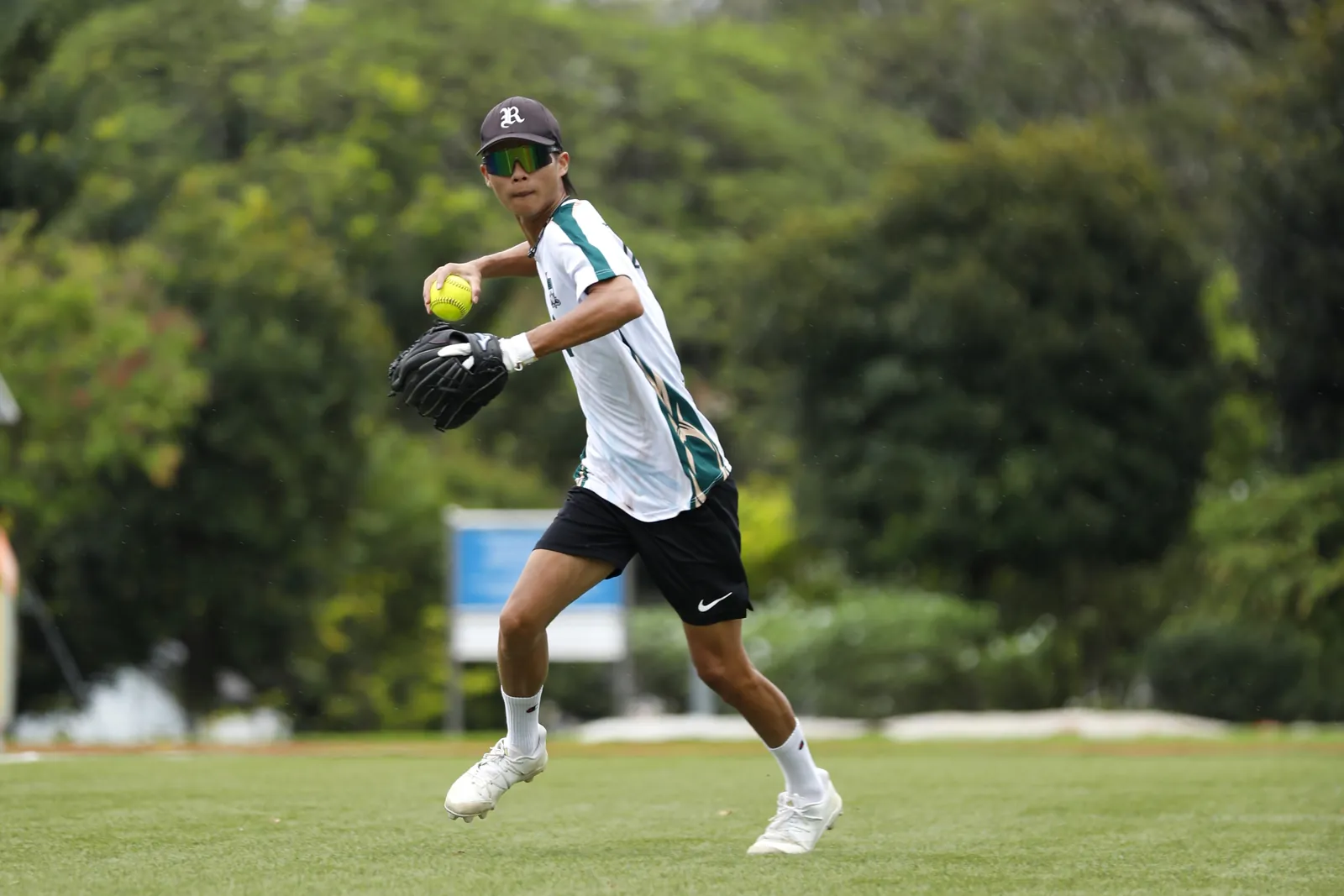 Softball player in a white and green jersey and reflective sunglasses preparing to throw a yellow ball.