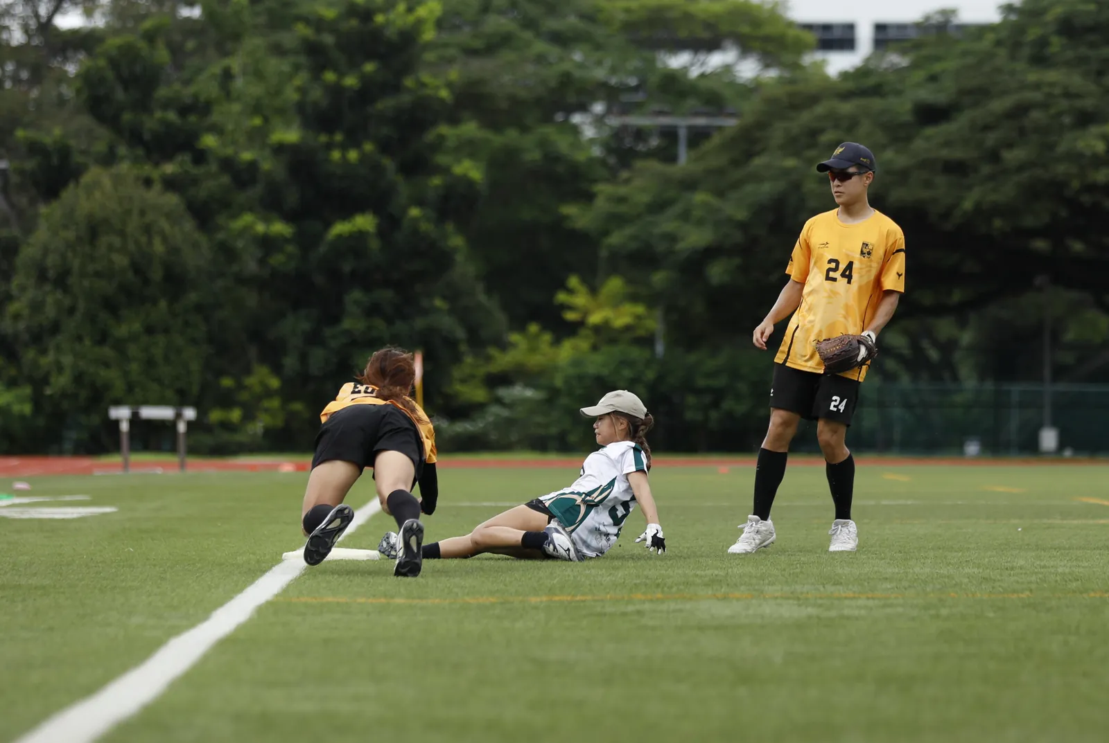 Softball runner in white and green sliding toward a base beside two fielders in yellow on a grass field.