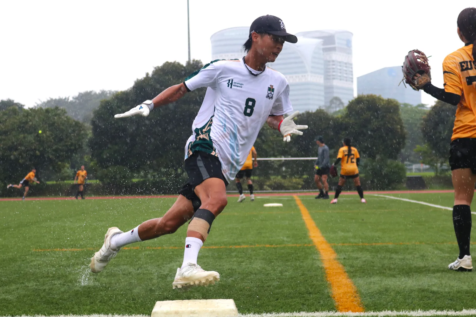 Softball runner wearing a white and green number 8 jersey rounding a base as rain falls across the field.